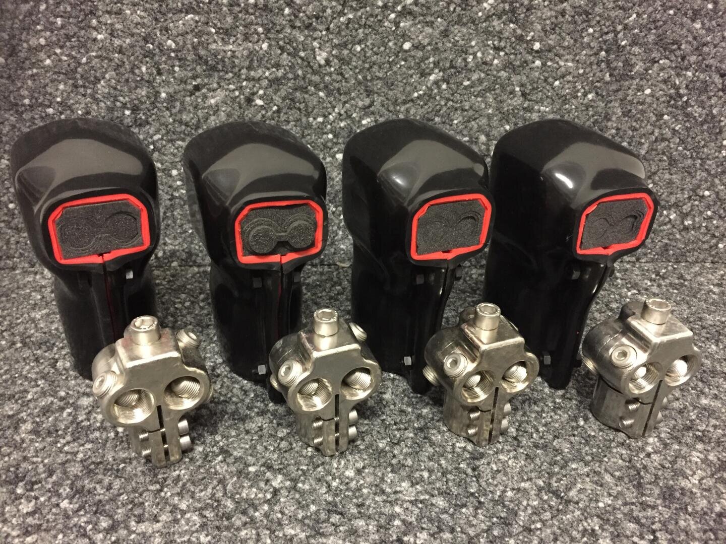 Vier schwarze Fahrradschalthebel mit roter Zierleiste und Schaumstoffeinlagen sind vor vier silbernen Fahrradkurbelarmen aus Metall auf einer gesprenkelten grauen Oberfläche ausgestellt.