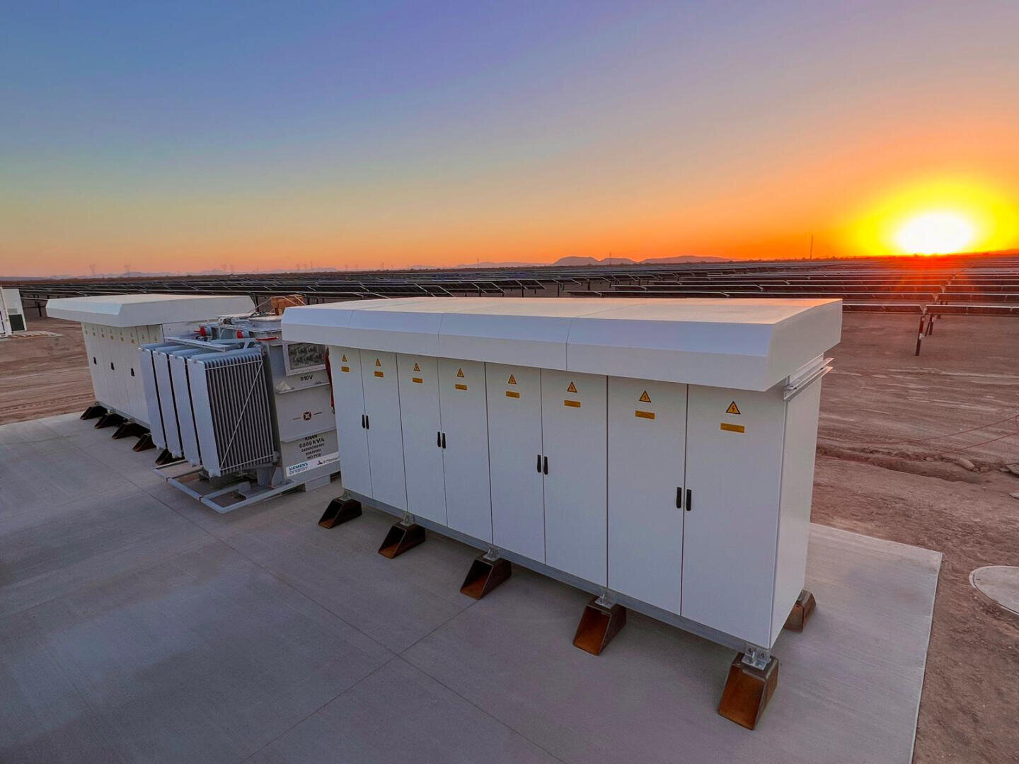 Rows of large white control cabinets and equipment stand on a concrete platform in a solar energy plant, with solar panels and a bright sunset in the background.
