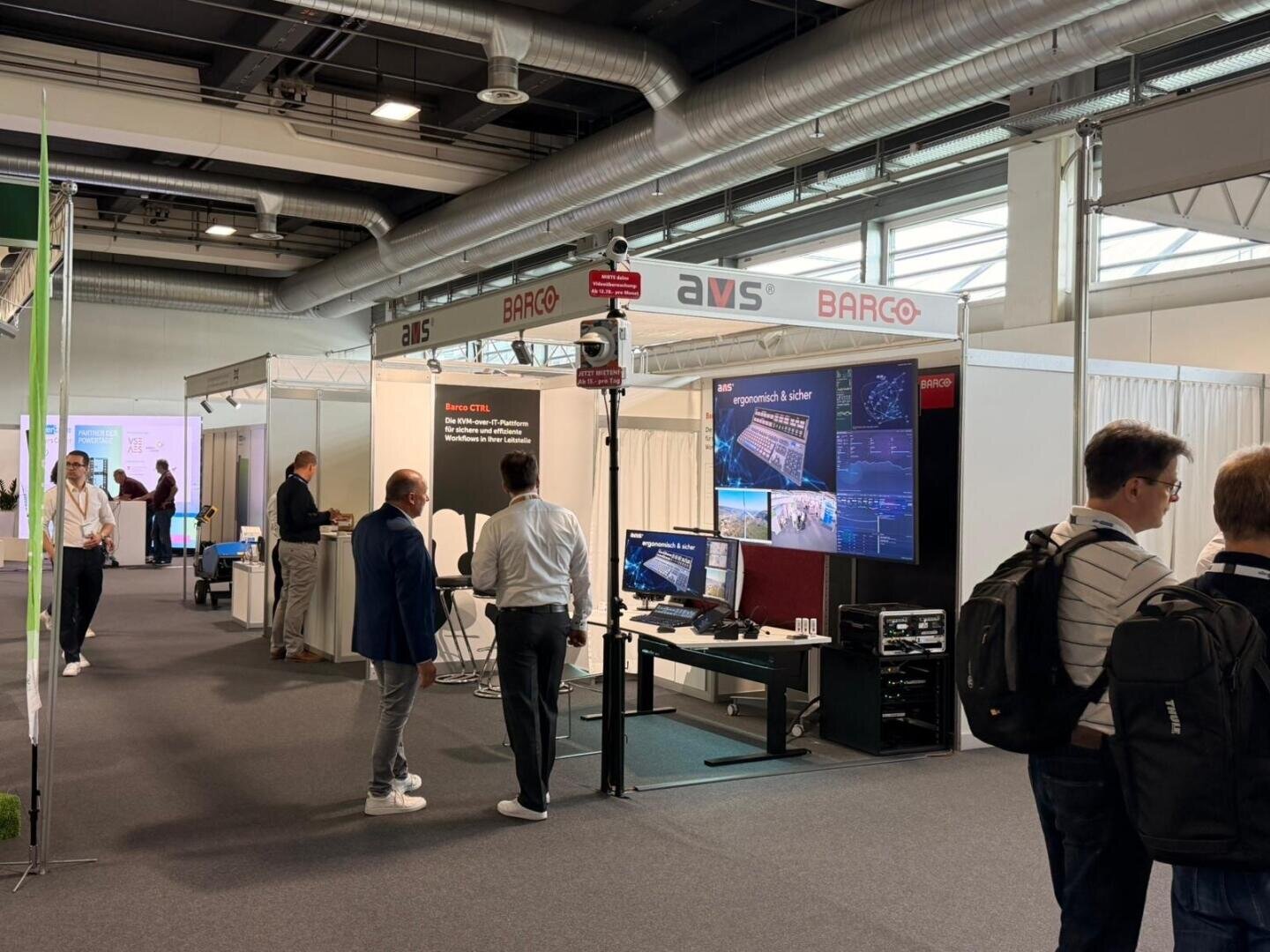 Ein Messestand, an dem Menschen stehen und sich unterhalten. Auf dem Stand sind mehrere Bildschirme, Geräte sowie Markenschilder von AVS und BARCO zu sehen. Im Hintergrund sind weitere Stände und Besucher zu erkennen.