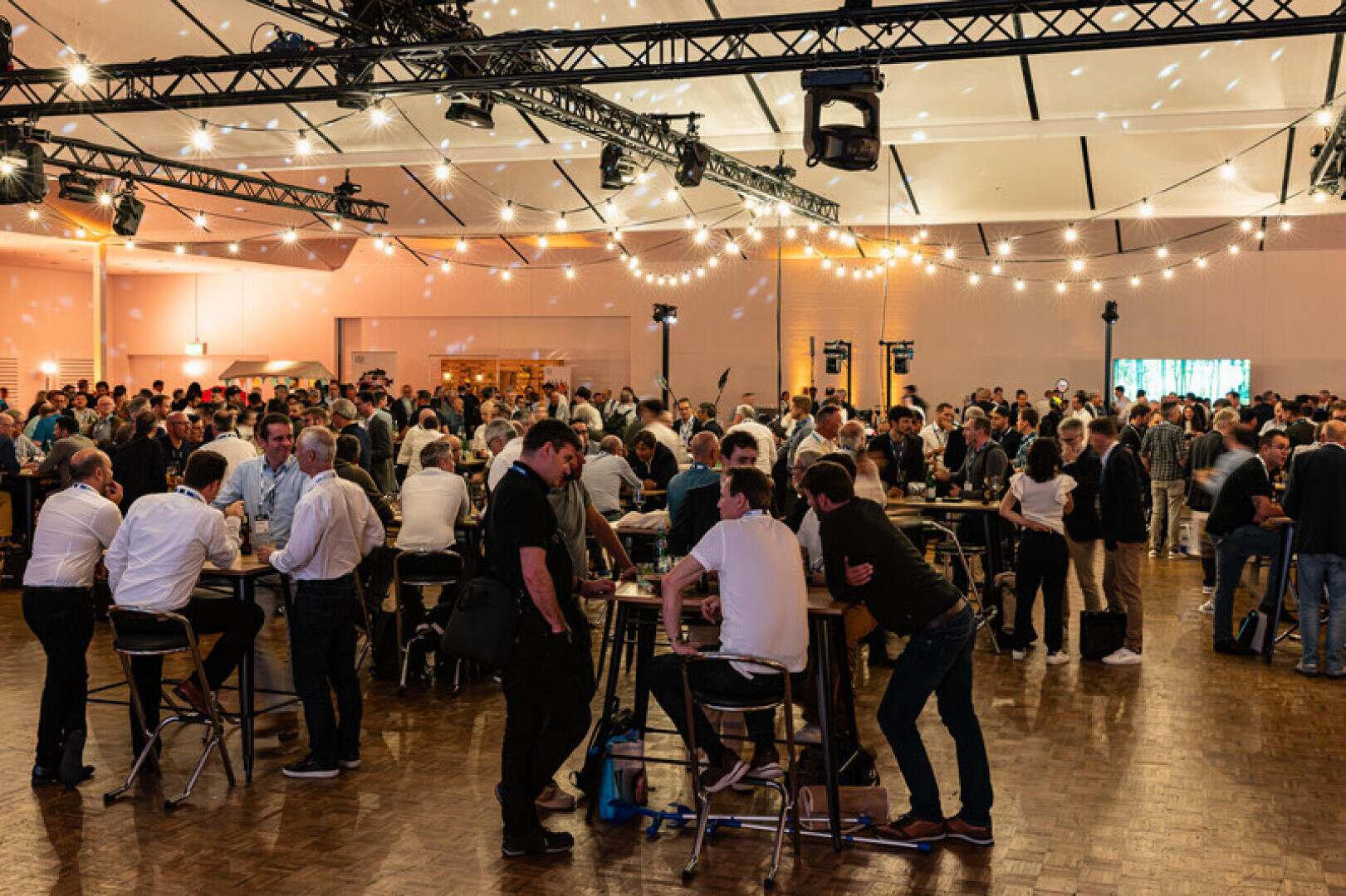 Eine große Gruppe von Menschen sitzt an Stehtischen bei einer Indoor-Veranstaltung mit Lichterketten und professioneller Beleuchtung über dem Kopf. Die Atmosphäre ist lebendig und festlich.