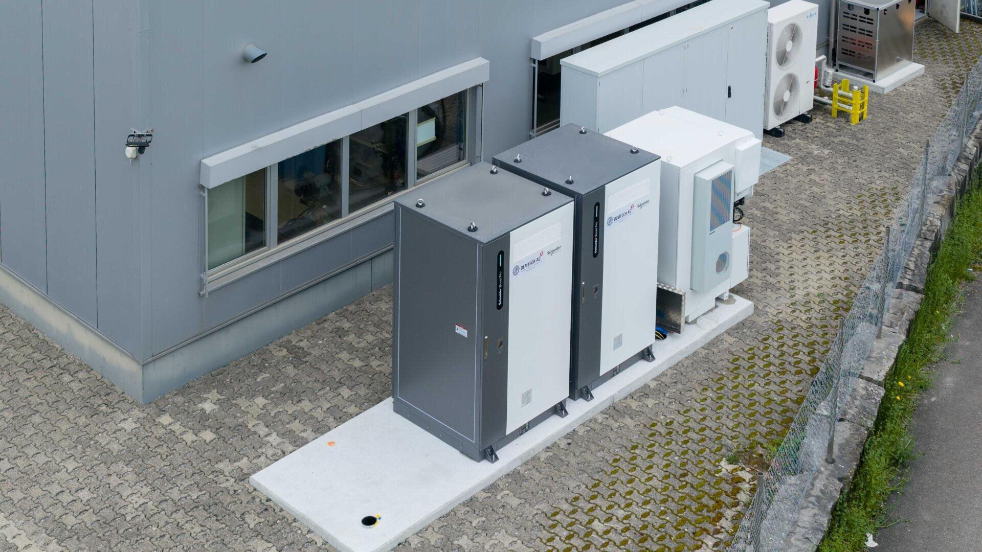 Zwei große grau-weiße Industrie-Schaltschränke sind außerhalb eines Gebäudes auf einer Betonfläche neben den HLK-Einheiten und einem Fenster installiert, mit einem gepflasterten Gehweg und einem Bordstein in der Nähe.