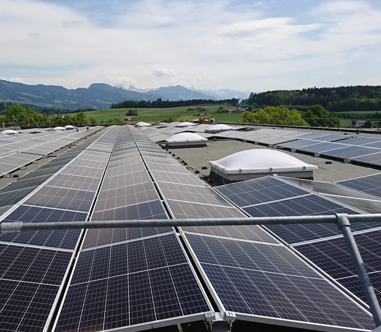 Reihen von Solarmodulen, die auf einem Dach installiert sind, mit grünen Feldern, Bäumen und entfernten Bergen unter einem teilweise bewölkten Himmel im Hintergrund.