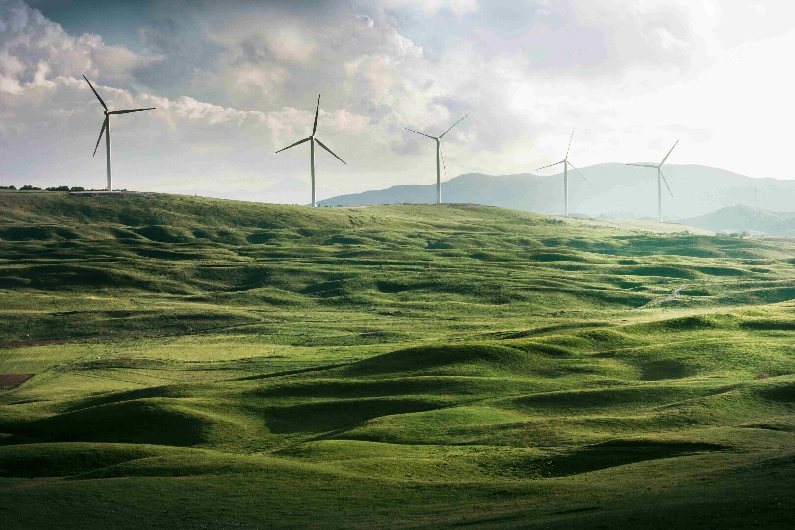 Windturbinen stehen auf einem entfernten Hügel über grünen Feldern bei teilweise bewölktem Himmel, im Hintergrund sind die Berge zu sehen.