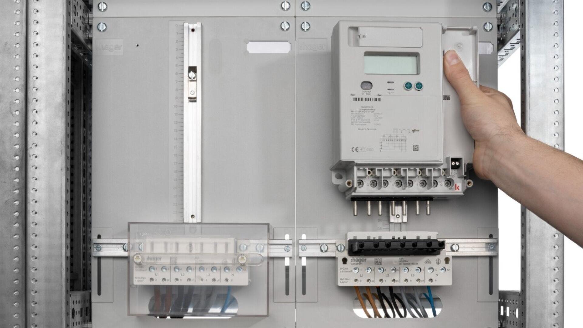 A person installs or removes a digital electricity meter inside an electrical panel where several wires and circuit breakers are visible. The meter has a display screen and buttons.