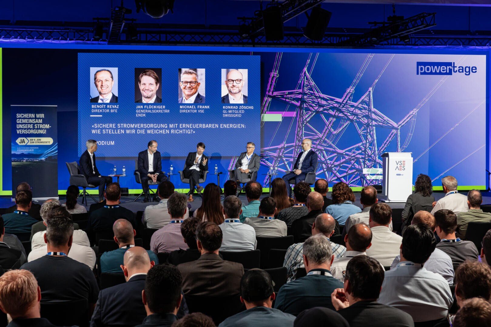 Podiumsdiskussion auf einer Konferenz, bei der fünf Redner auf der Bühne vor einem großen Publikum sitzen. Hinter ihnen befindet sich ein Bildschirm, auf dem ihre Namen, Fotos und das Thema nachhaltige Energie zu sehen sind.