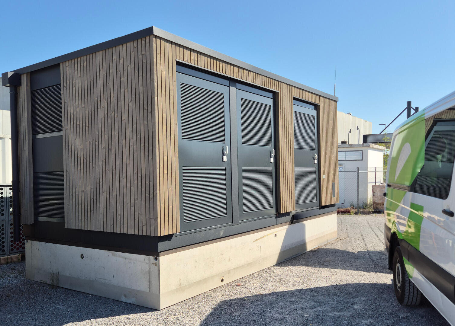 Eine moderne Holztrafostation steht auf einem Betonfundament unter freiem Himmel, neben einem weiß-grünen Eniwa-Lieferwagen.