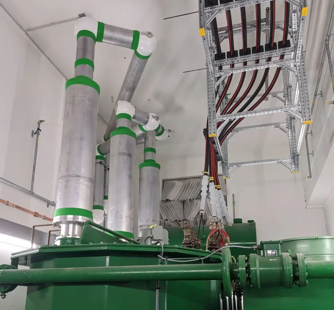 Große silberne Rohre mit grünen Bändern sind mit grünen Industriemaschinen in einem sauberen, weißen Raum verbunden. Über dem Kopf trägt eine metallene Kabelrinne mehrere rote und schwarze Kabel.