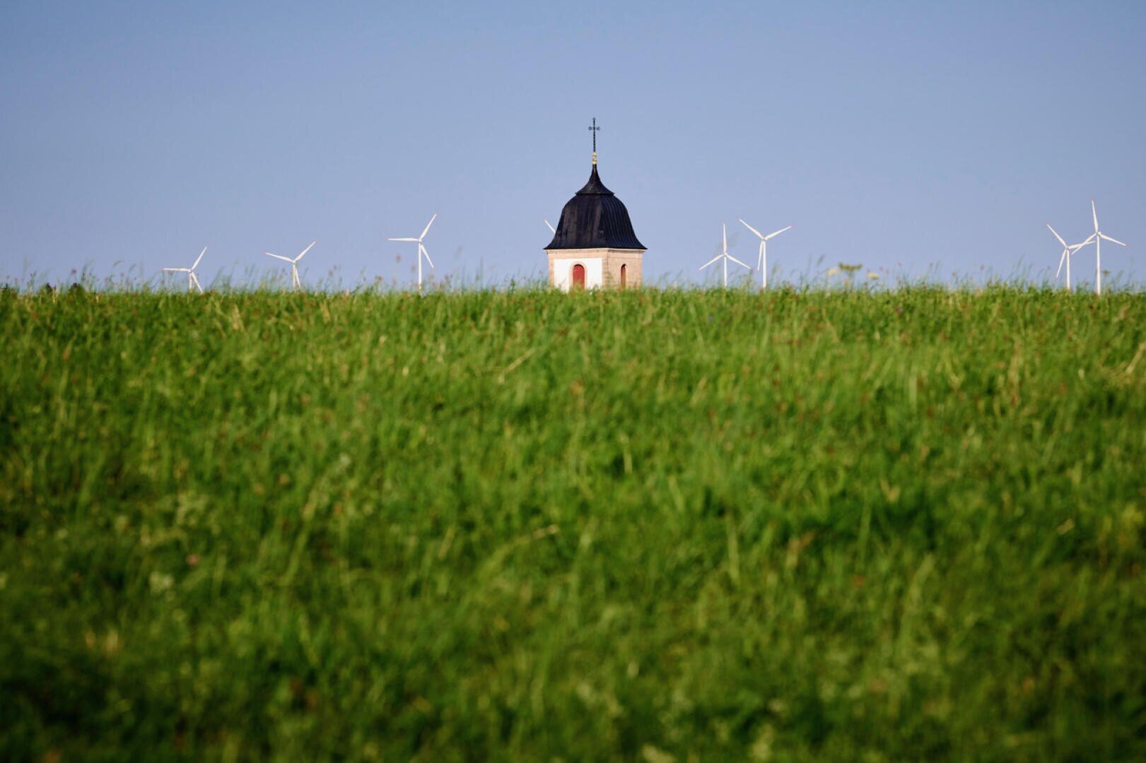 Ein Kirchturm mit einem Kreuz erhebt sich über einem üppigen grünen Feld, im Hintergrund sind mehrere Windturbinen vor einem klaren blauen Himmel zu sehen.