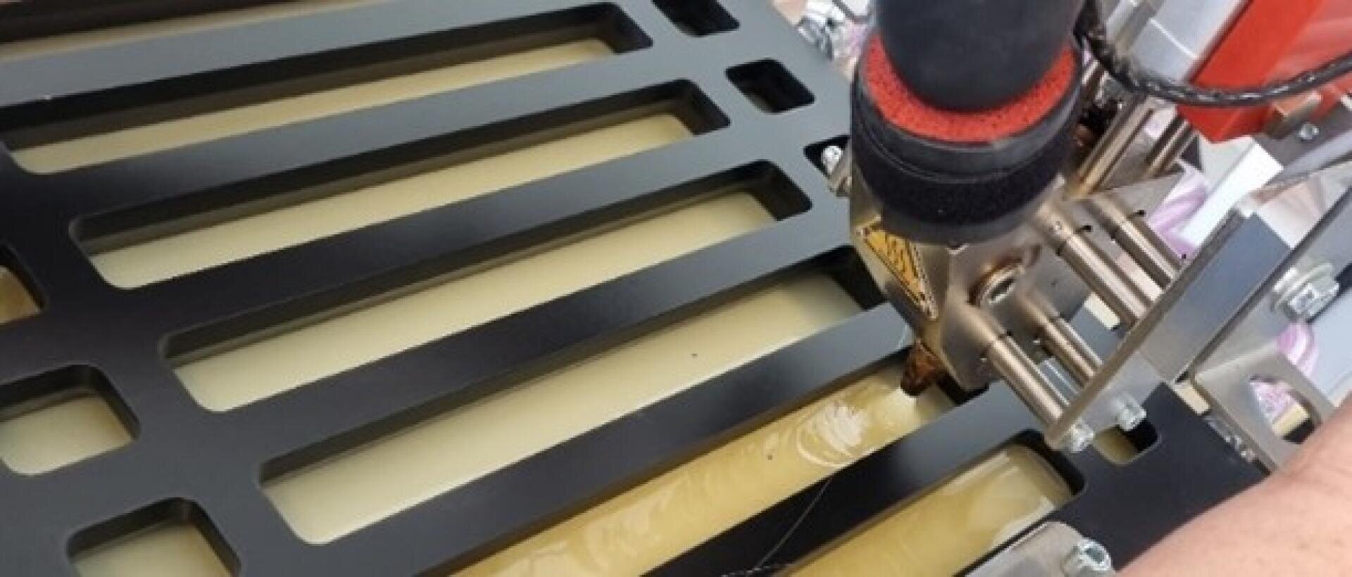 Nahaufnahme einer Maschine, die durch einen geschlitzten Metallrahmen Material auf eine flache Oberfläche extrudiert, möglicherweise während eines Herstellungs- oder 3D-Druckverfahrens.