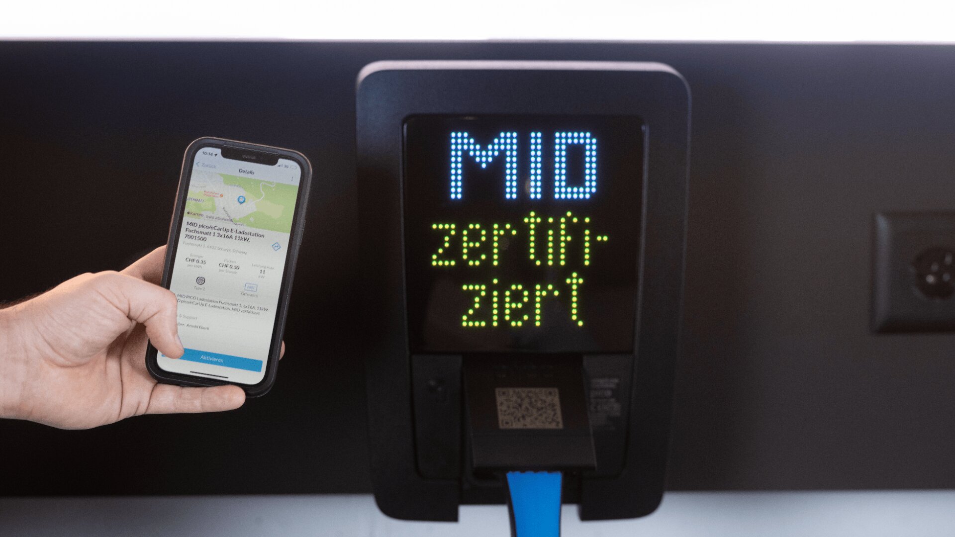 Eine Hand hält ein Smartphone mit einer App vor einem digitalen Bildschirm, auf dem die Worte MID zertifiziert mit einem blauen Kabel und einem QR-Code unterhalb des Bildschirms zu sehen sind.