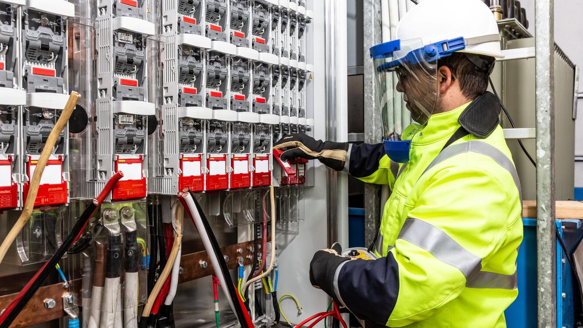 Ein Elektriker, der einen Schutzhelm, einen Gesichtsschutz und eine Warnweste trägt, arbeitet an einer elektrischen Schalttafel mit mehreren Leistungsschaltern und roten Schildern in einer Industrieumgebung.