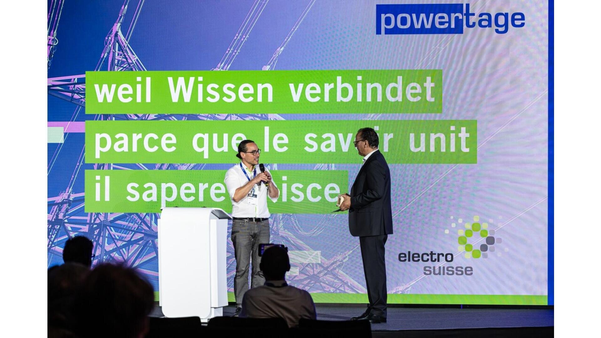 Two men stand on a stage in front of a screen displaying “weil Wissen verbindet, parce que le savoir unit, il sapere unisce” and logos for powertage and electro suisse. One man holds a microphone and speaks to the other.
