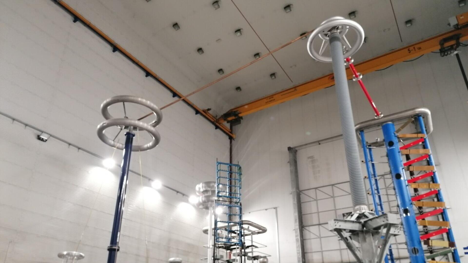 Großes Indoor-Hochspannungsprüflabor mit hohen elektrischen Geräten, darunter Metallringe und Isolatoren, unter einer hohen Decke mit einem gelben Brückenkran. Helle Lichter erhellen die industrielle Umgebung.
