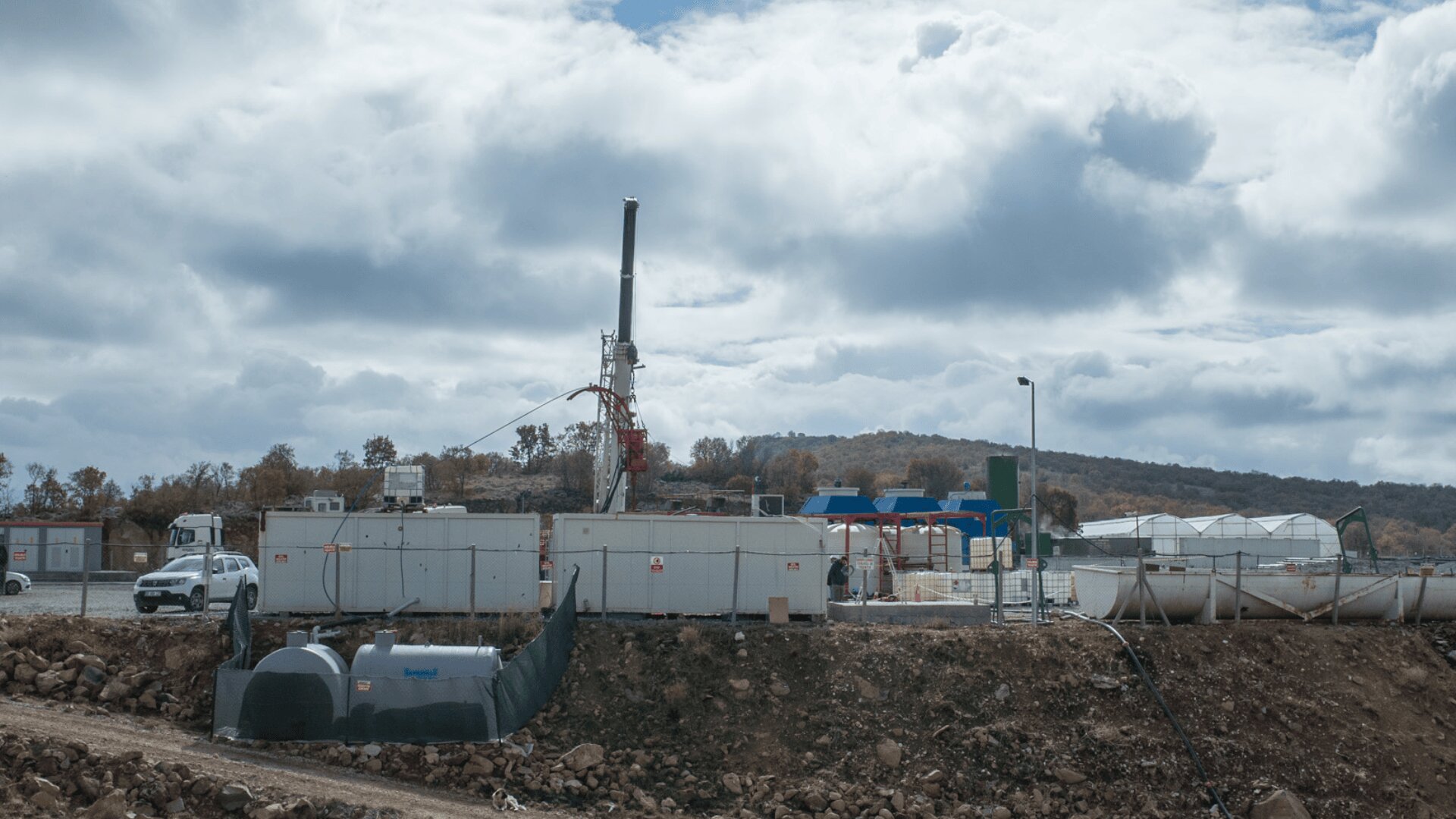 Eine Bohrstelle mit Industrieanlagen, Containern und einem Bohrturm bei bewölktem Himmel. Ein weißes Fahrzeug ist in der Nähe geparkt, und das Gelände ist von einem Zaun und felsigem Boden umgeben. Im Hintergrund sind Hügel mit Herbstbäumen zu sehen.