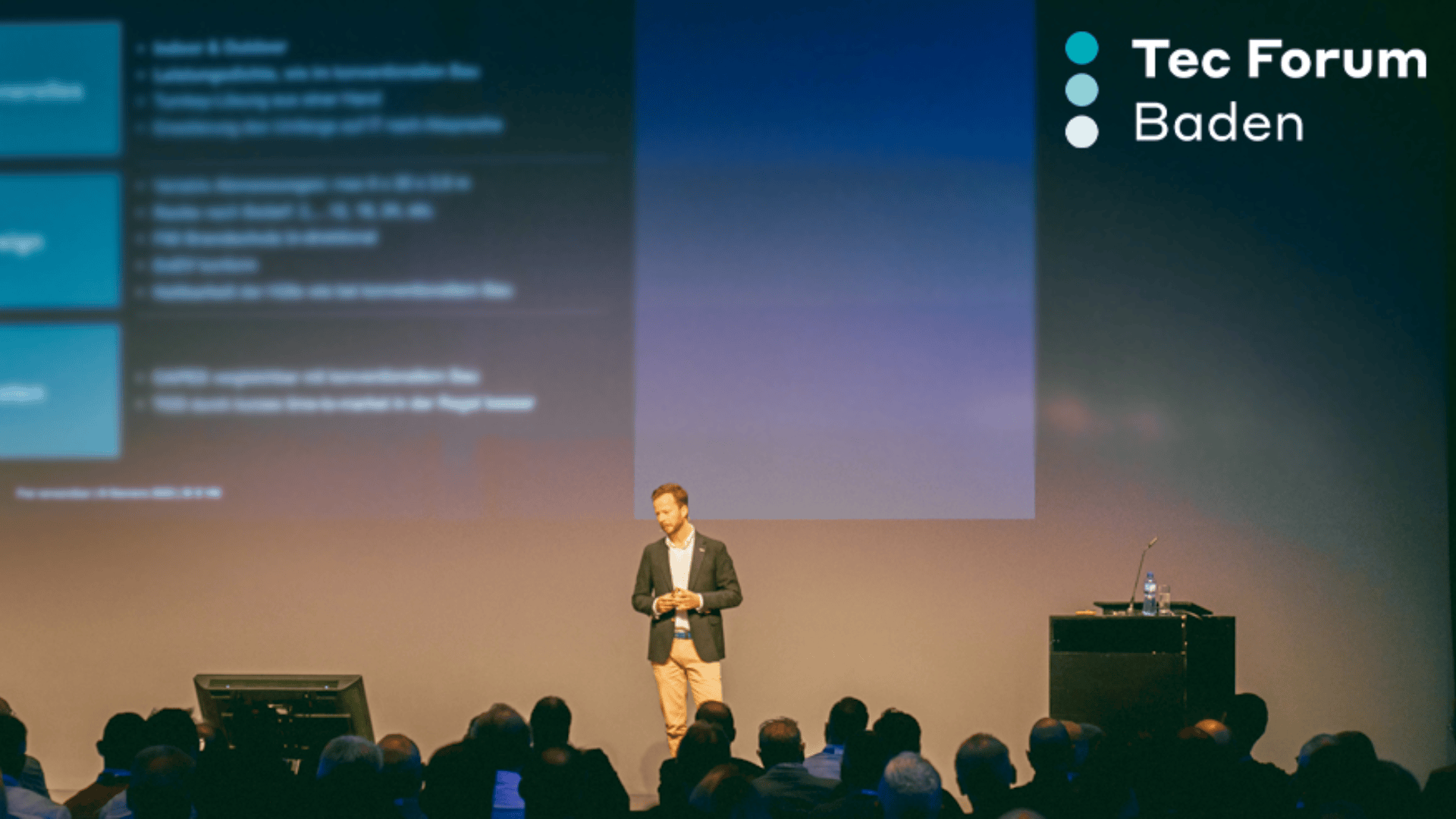 A man in a blazer stands on stage presenting to an audience at Tec Forum Baden; slides are projected behind him, and the event logo is visible in the top right corner.
