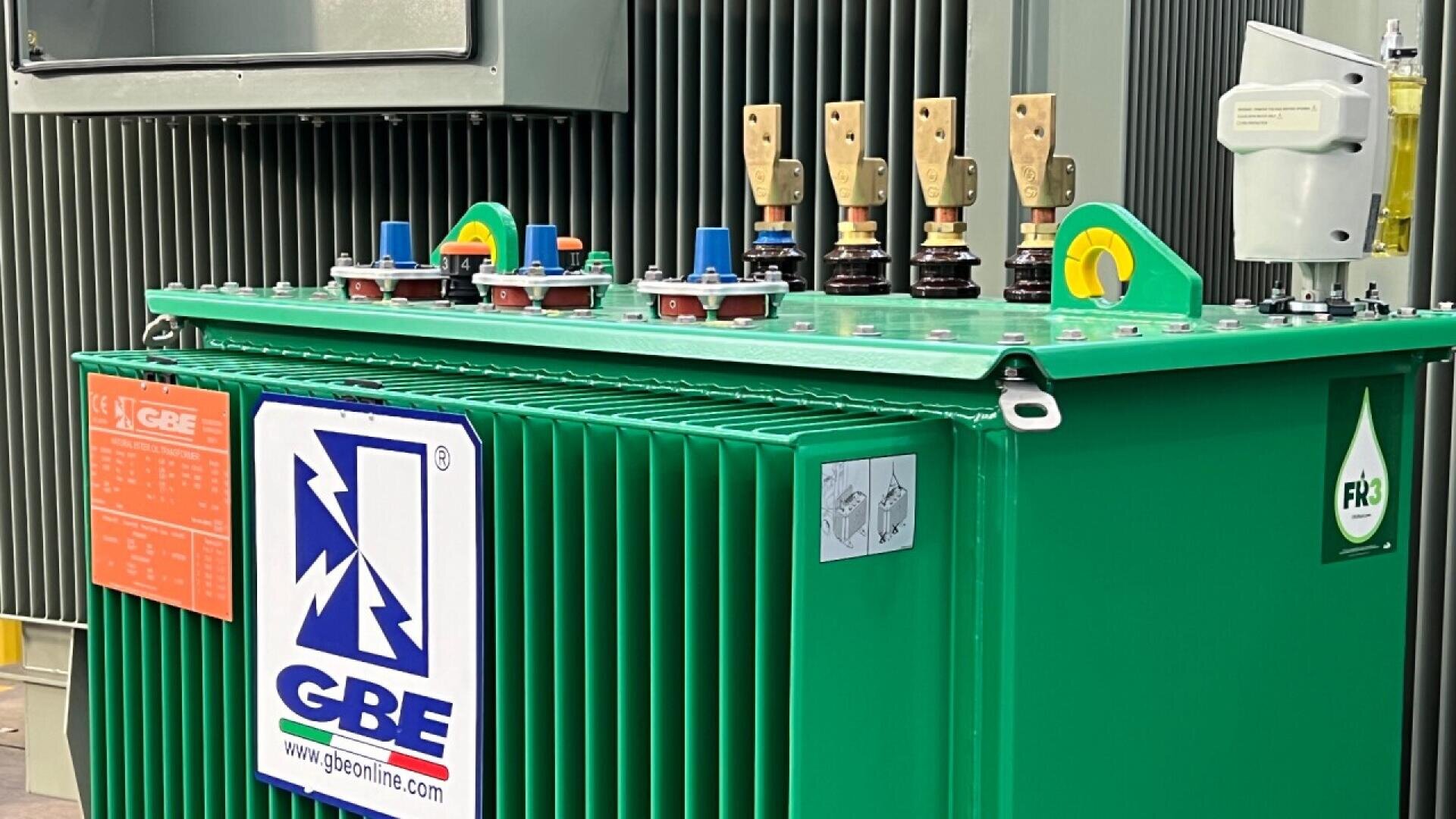 Ein grüner elektrischer Transformator mit beschrifteten Bedienelementen und Schaltern an der Oberseite, mit dem GBE-Firmenlogo und der Website an der Seite; im Hintergrund sind Metallrippen zur Kühlung zu sehen.