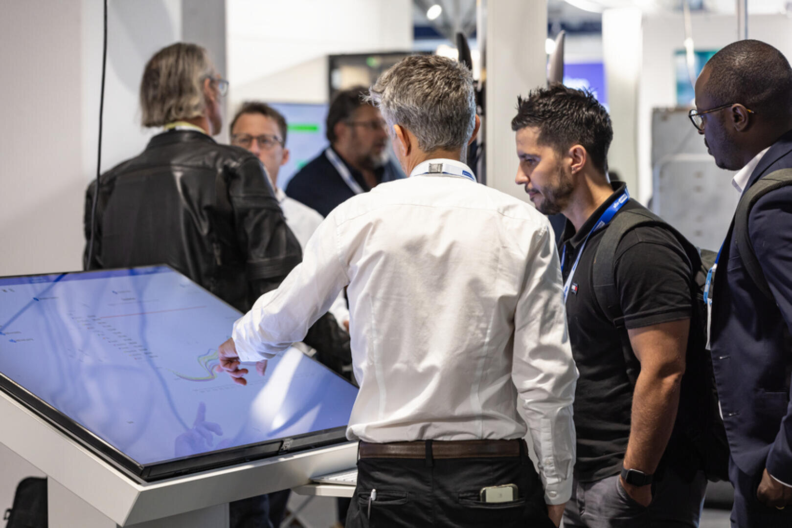 A group of men gathered around a large touchscreen display at a technology event, with one man pointing at the screen while others watch attentively.