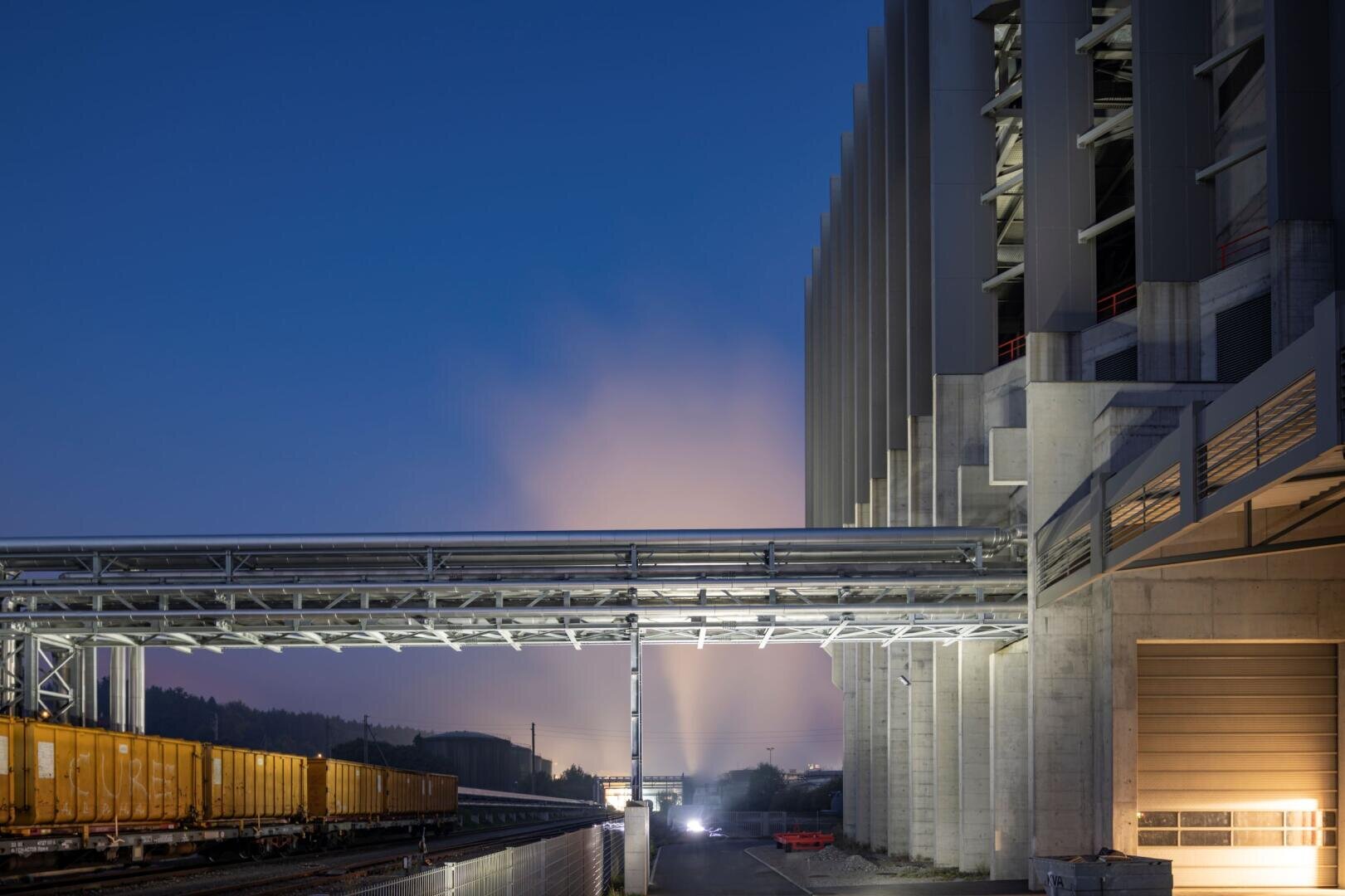 Eine moderne Industrieanlage in der Abenddämmerung mit großen Betonstrukturen, Metallrohren und gelben Güterzugwaggons auf den Gleisen. Helle Lichter erhellen die Szene, und im Hintergrund steigt Nebel oder Rauch auf.