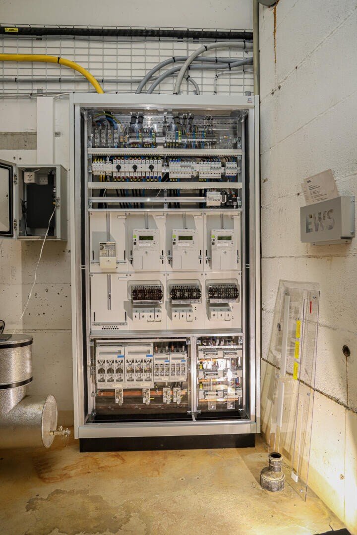 Eine große elektrische Schalttafel mit mehreren Schaltern, Zählern, Kabeln und Leistungsschaltern ist an einer Wand in einer Industrieanlage montiert, über der und um die herum Rohre und Kabel verlaufen.