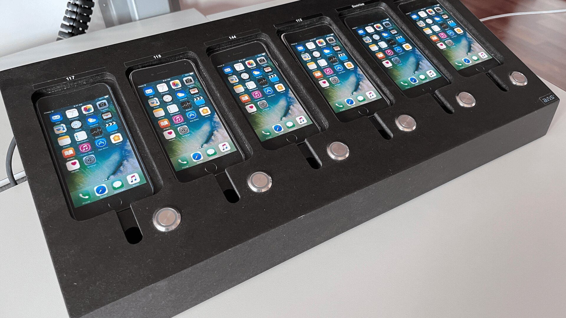 Sechs iPhones werden in einer schwarzen Schaumstoffschale ausgestellt, jedes mit eingeschaltetem Bildschirm, auf dem der Startbildschirm mit App-Symbolen angezeigt wird. Jedes Telefon befindet sich in seinem eigenen Steckplatz über einer runden silbernen Taste.