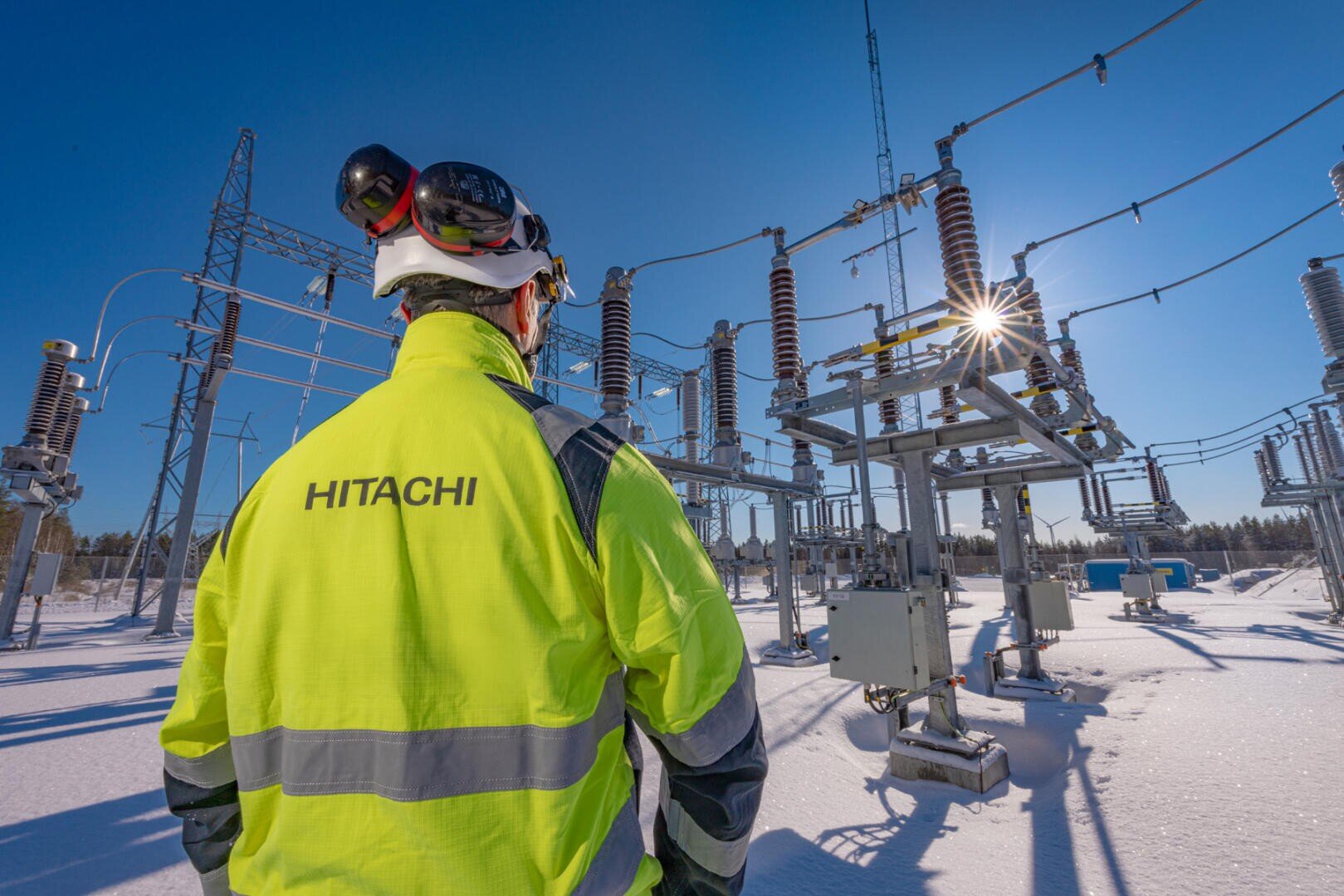 Ein Arbeiter in einer Warnweste mit der Aufschrift HITACHI steht in einem verschneiten Umspannwerk unter strahlend blauem Himmel, umgeben von Stromleitungen und Geräten.