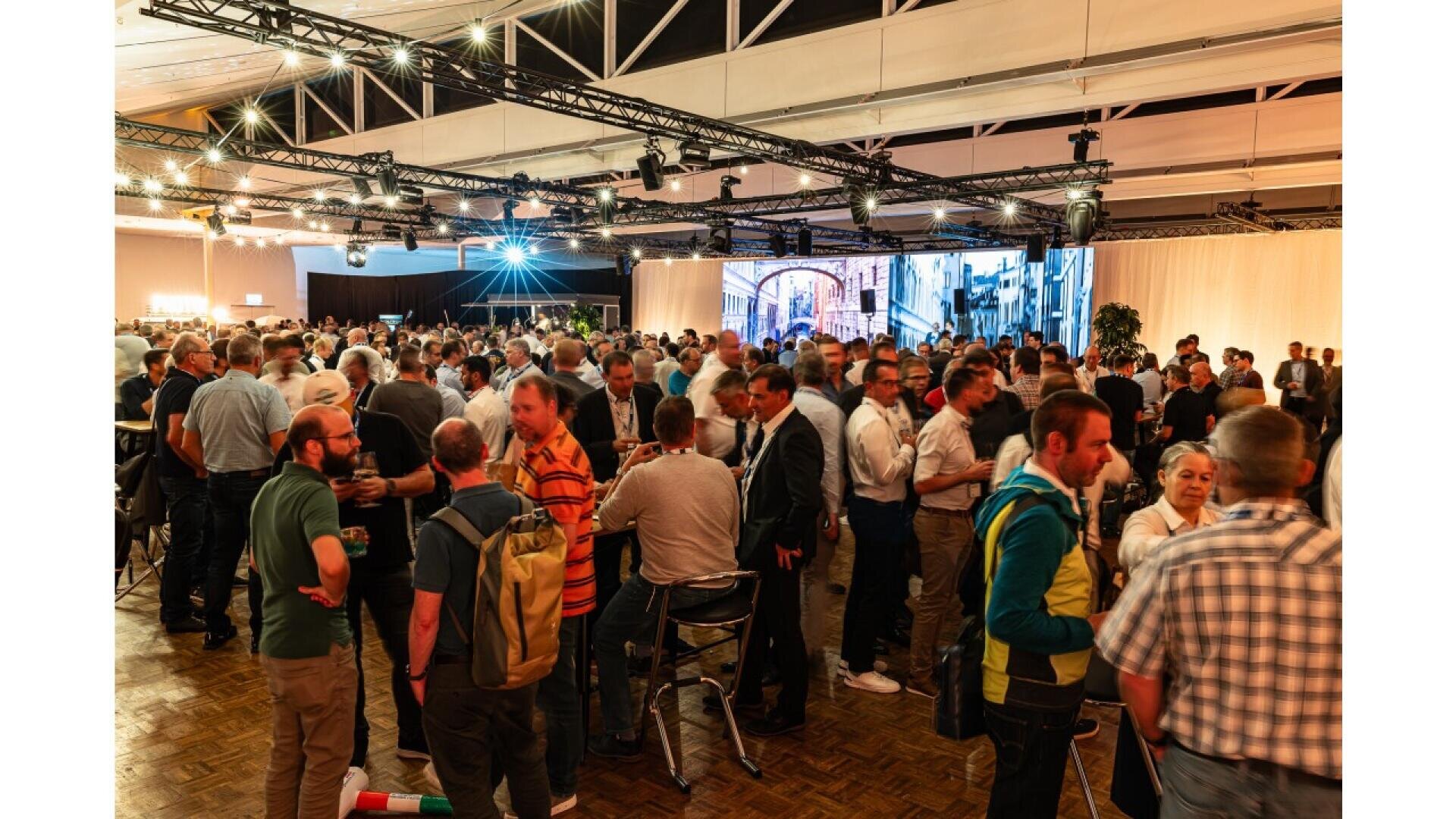 A large group of people socializing at an indoor event or conference, with various small groups talking, some holding drinks, under bright lights and a high ceiling.