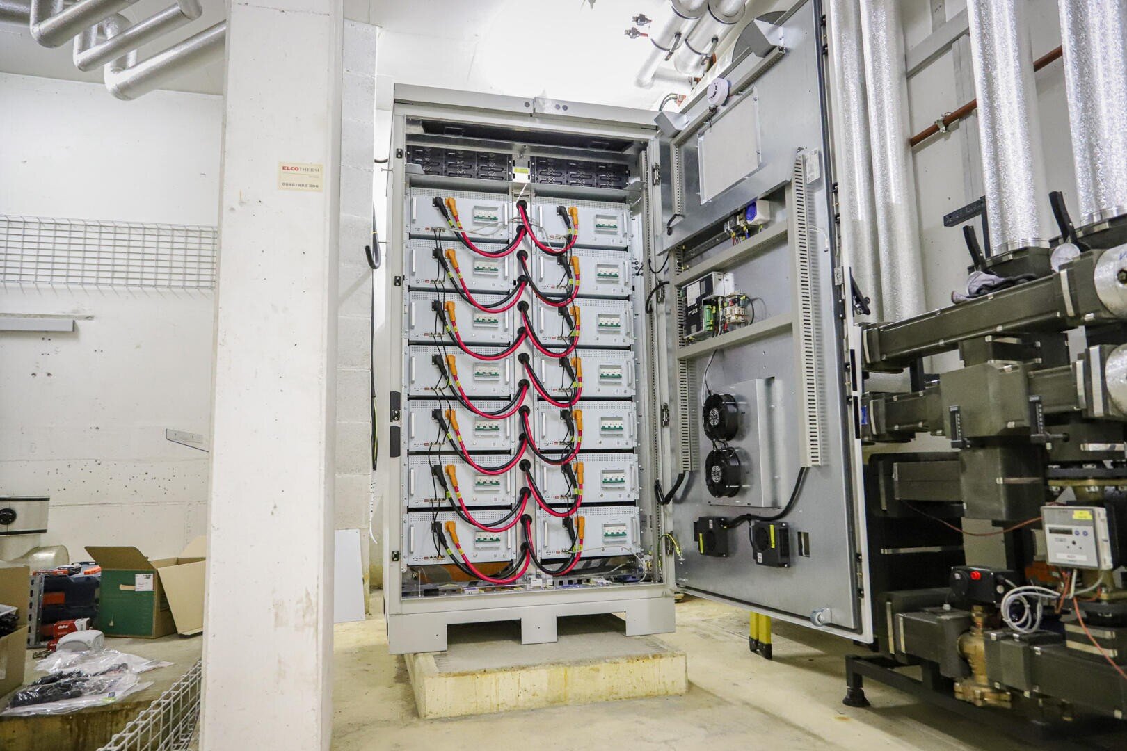 Ein offener Industrie-Elektroschrank zeigt mehrere gestapelte Batterieeinheiten, die mit roten und schwarzen Kabeln verbunden sind, umgeben von Rohren, Maschinen und Werkzeugen in einem Versorgungsraum.