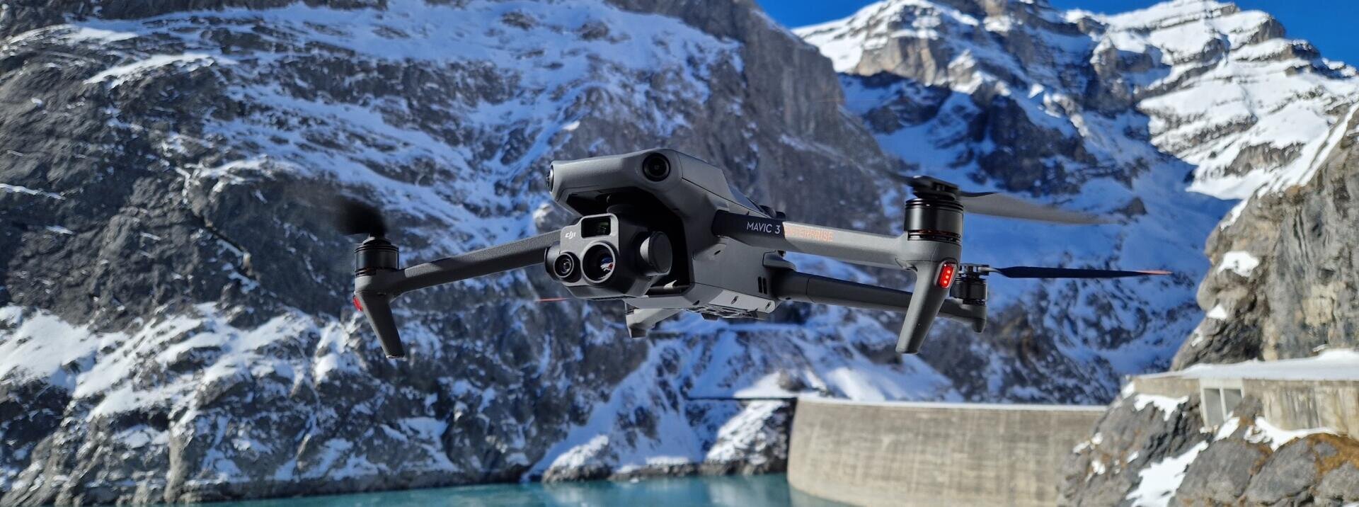 A black drone with cameras flies in front of snow-covered mountains and a blue lake, with part of a dam visible in the background on a clear day.