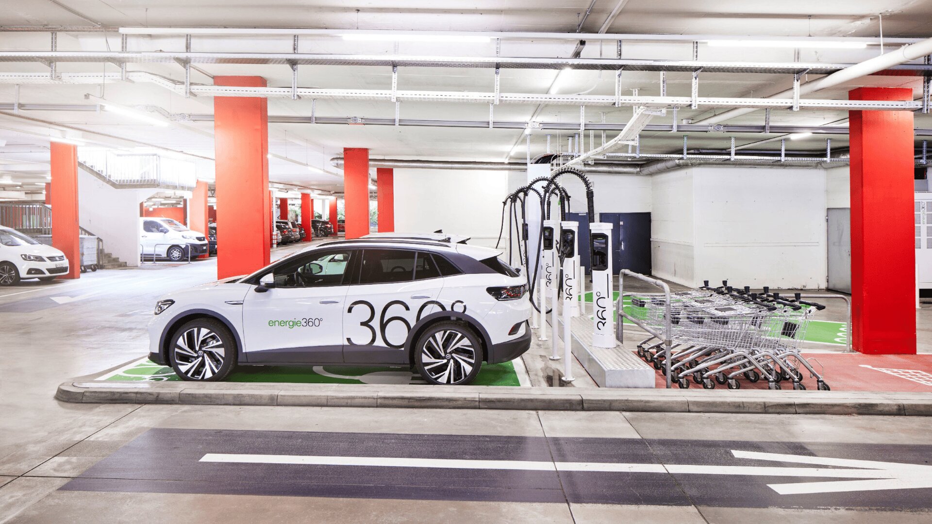 Ein Elektroauto parkt und lädt an einer Indoor-Ladestation neben einer Reihe von Einkaufswagen in einem Parkhaus mit roten Säulen.