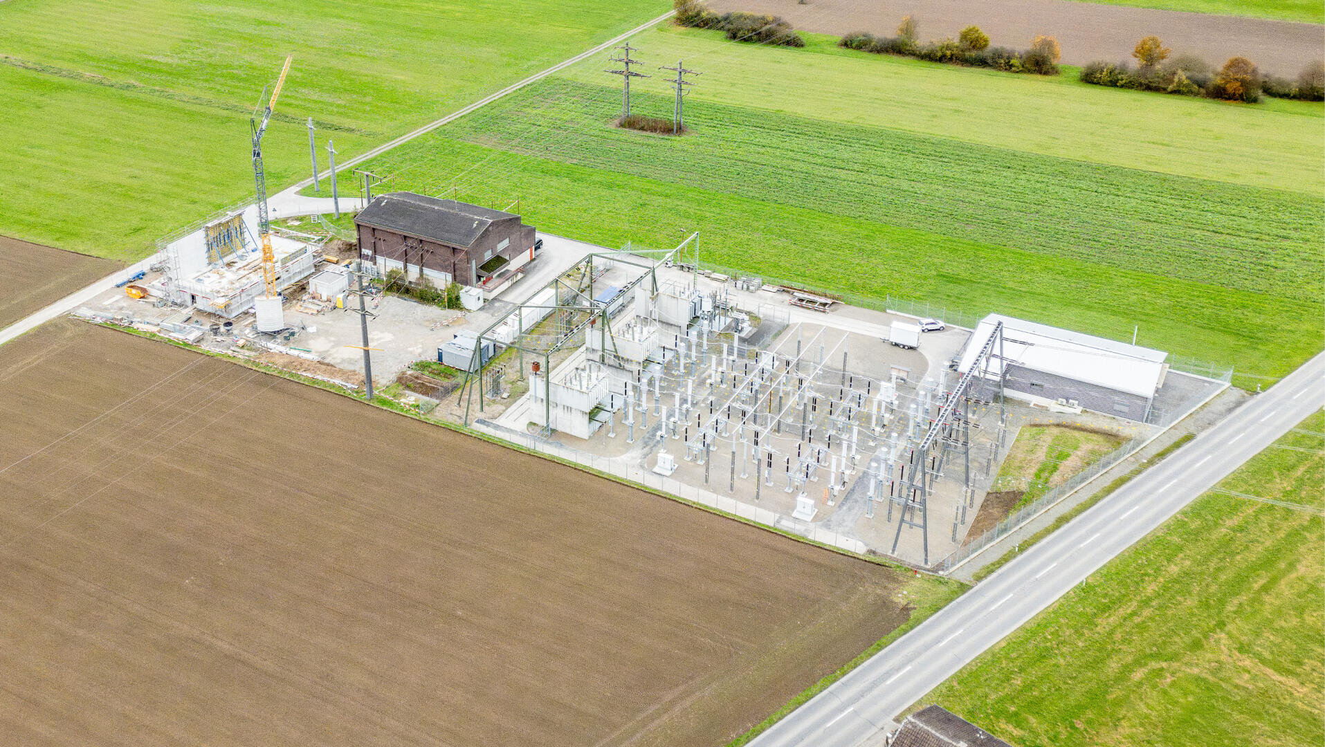 Aerial view of a substation surrounded by green fields and farmland, with electricity pylons, transformers and fences along a nearby road.