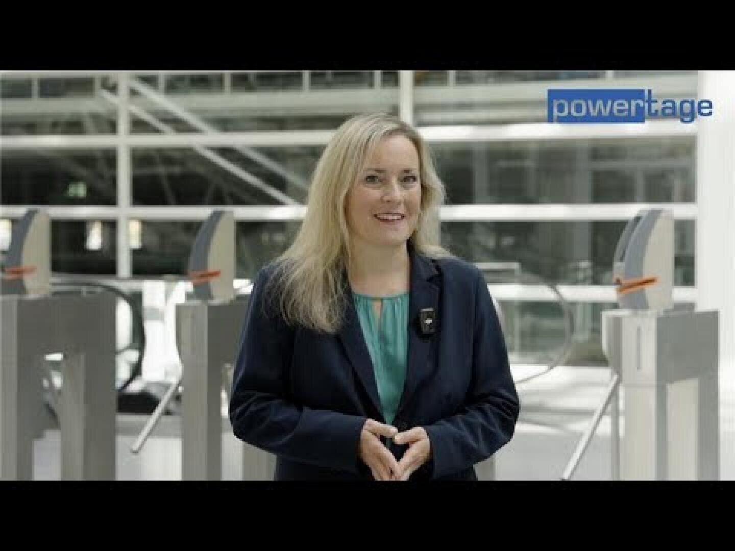Eine Frau mit langen blonden Haaren, die ein blaugrünes Oberteil und einen marineblauen Blazer trägt, steht in einem Innenraum vor einem modernen Sicherheitstor. In der rechten oberen Ecke erscheint das Wort powertage.