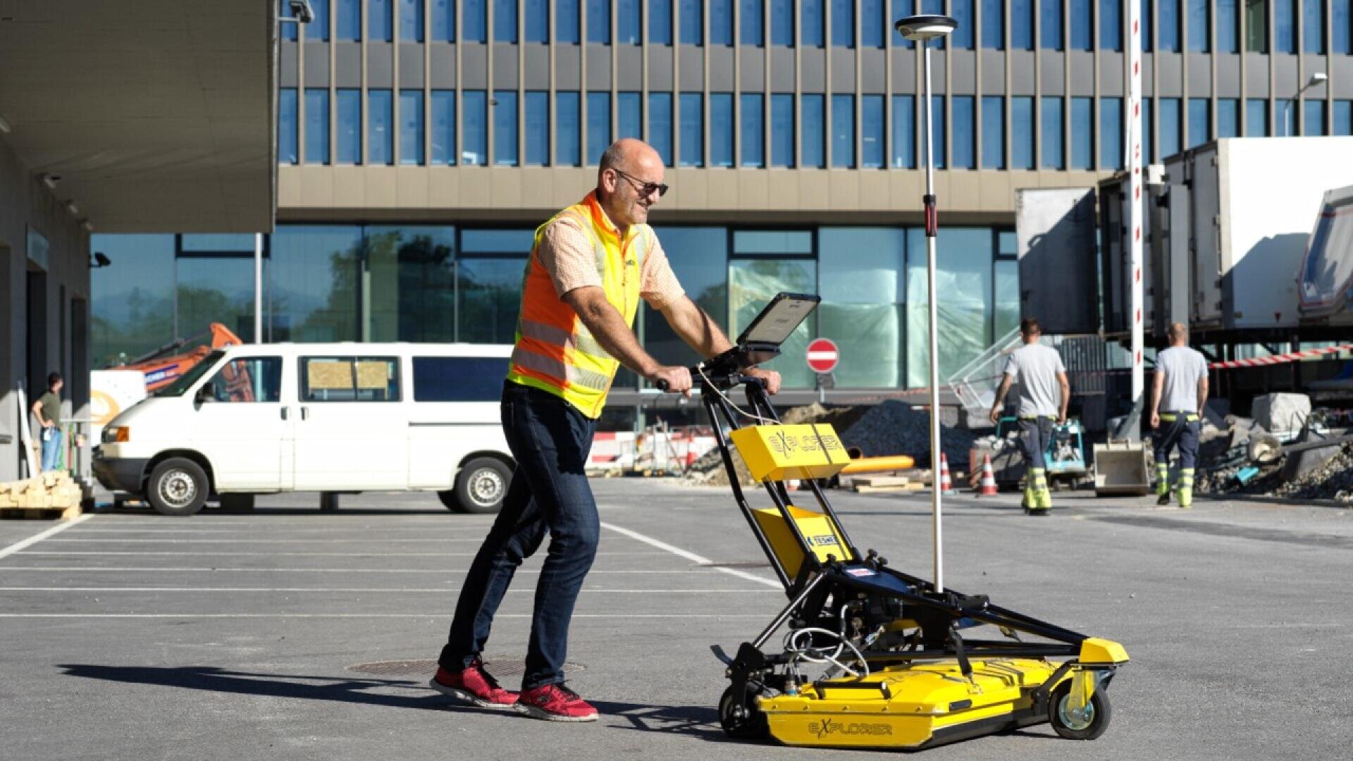 A man in a safety vest and sunglasses uses a large yellow ground-penetrating radar device on wheels in a parking lot near a construction site, with workers and vehicles in the background.