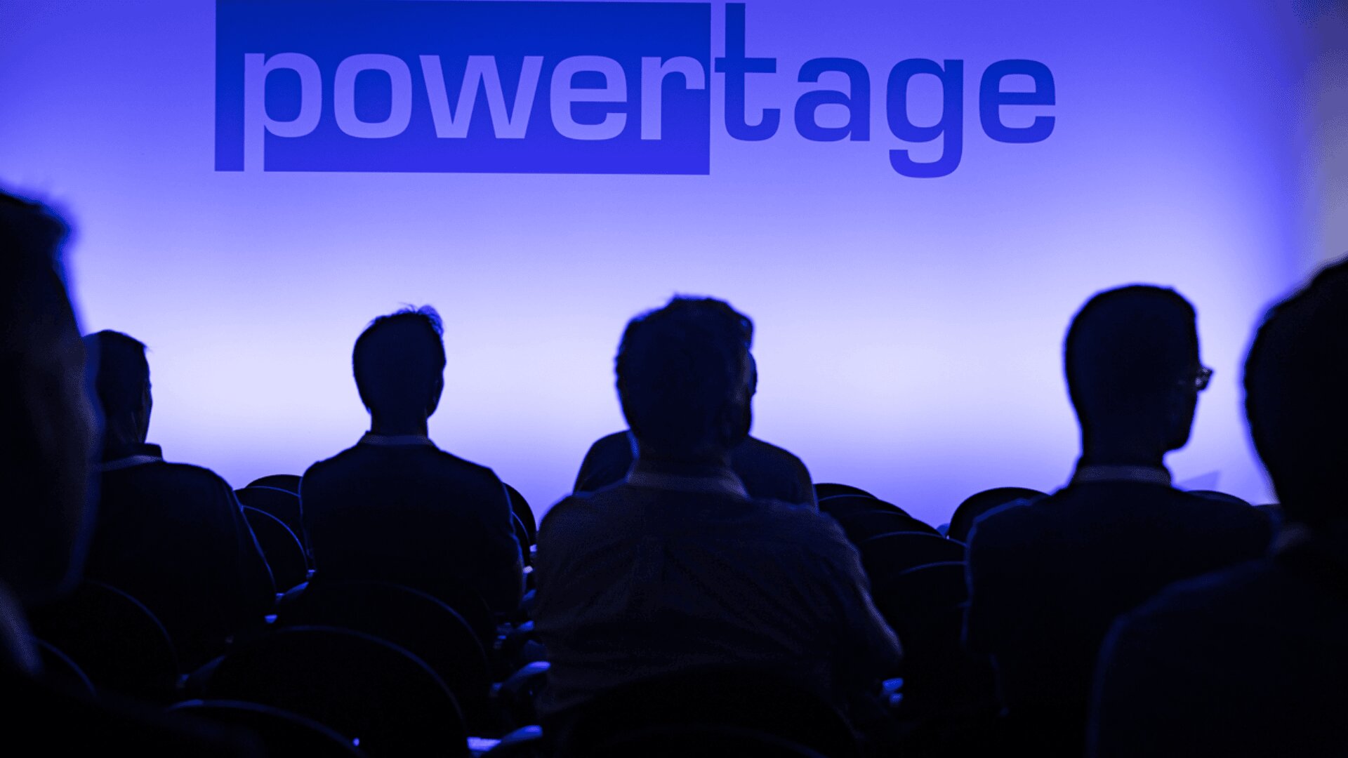 Silhouetten von Menschen, die im Publikum sitzen und auf einen Bildschirm oder eine Wand blicken, auf der das Wort powertage in blauem Licht leuchtet.