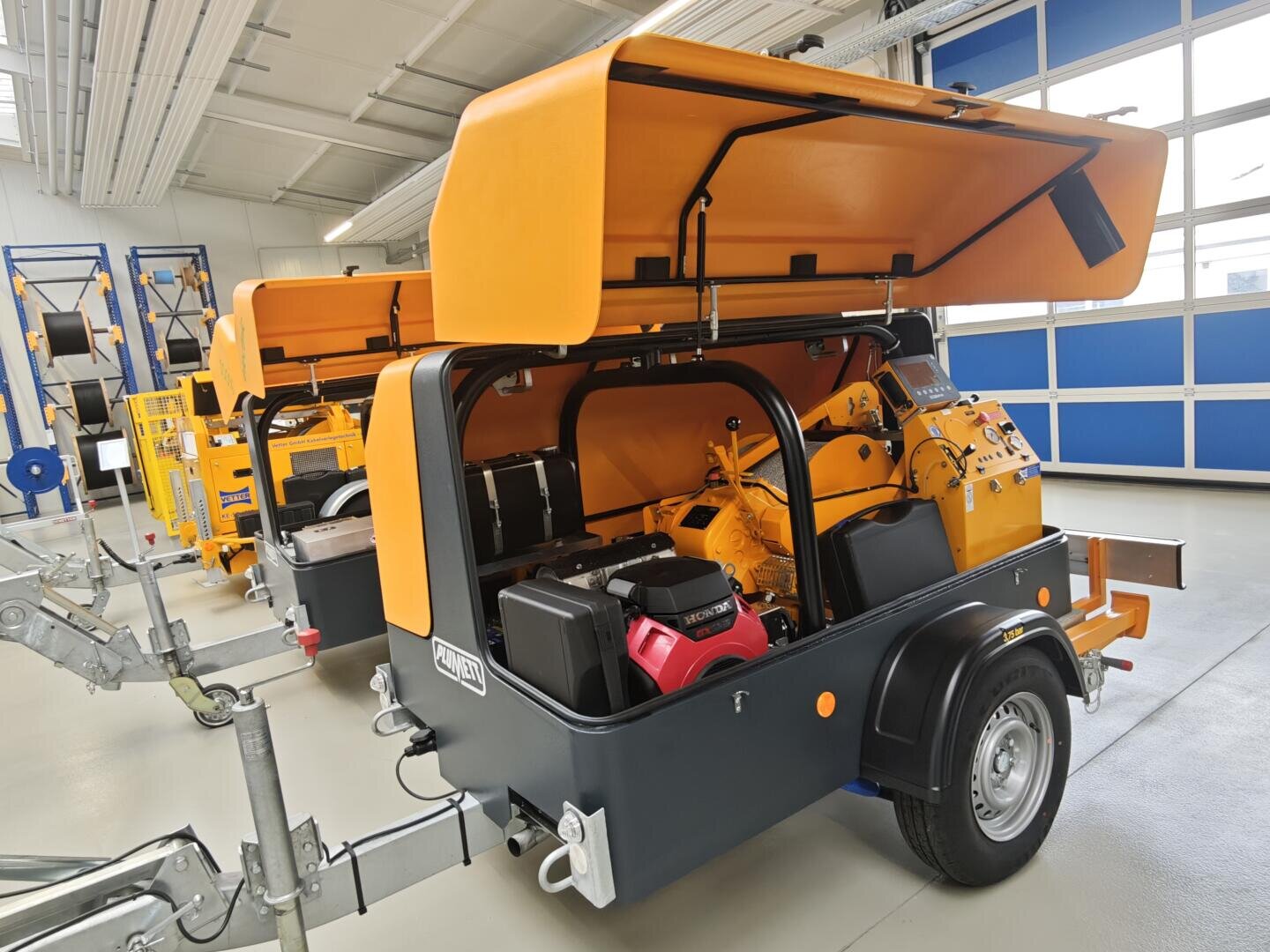 In einer sauberen, gut beleuchteten Werkstatt mit blauen und weißen Wänden und großen Fenstern stehen zwei mobile Industriemaschinen, deren orangefarbene Abdeckhauben geöffnet sind und den Blick auf mechanische Komponenten im Inneren freigeben.