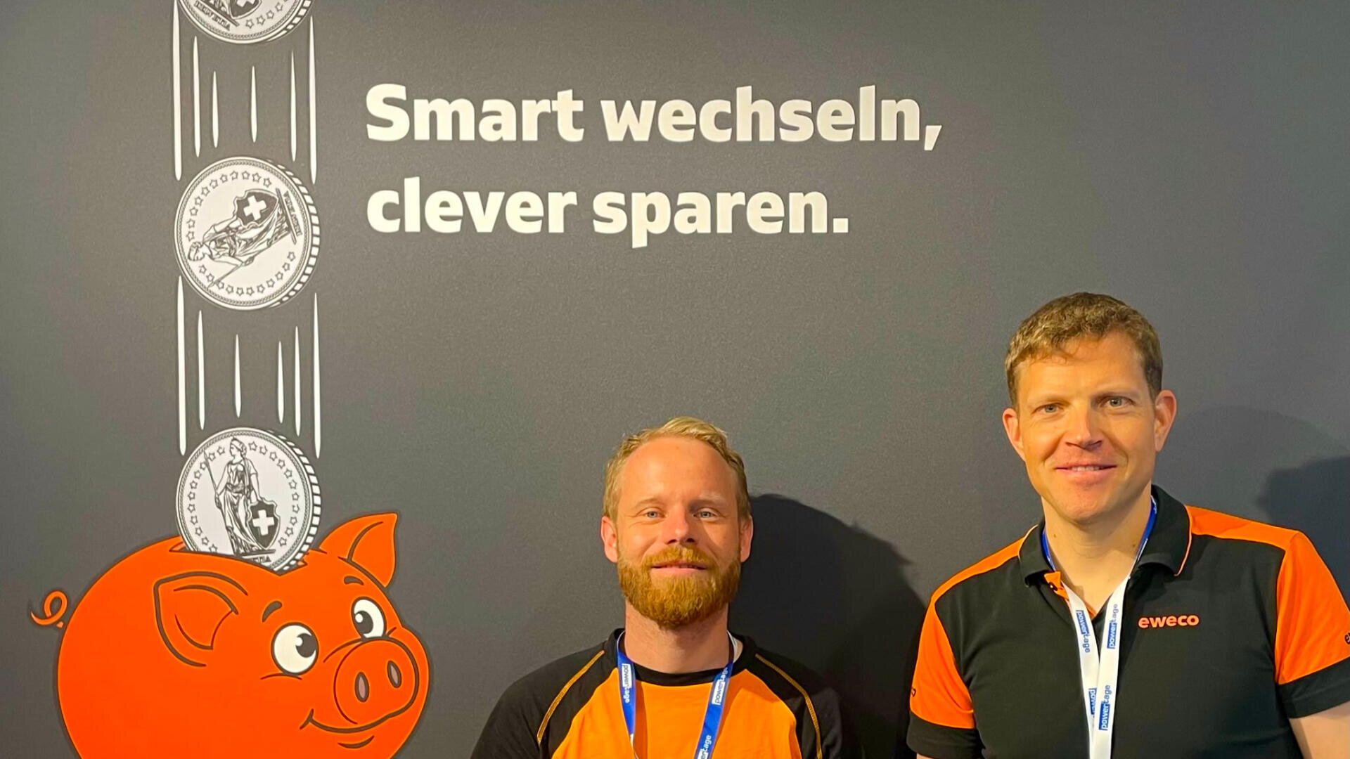 Zwei Männer in schwarzen und orangefarbenen Hemden stehen vor einer Wand, auf der der deutsche Text „Smart wechseln, clever sparen“ zu lesen ist, sowie ein Cartoon-Sparschwein, in das Münzen hineinfallen.
