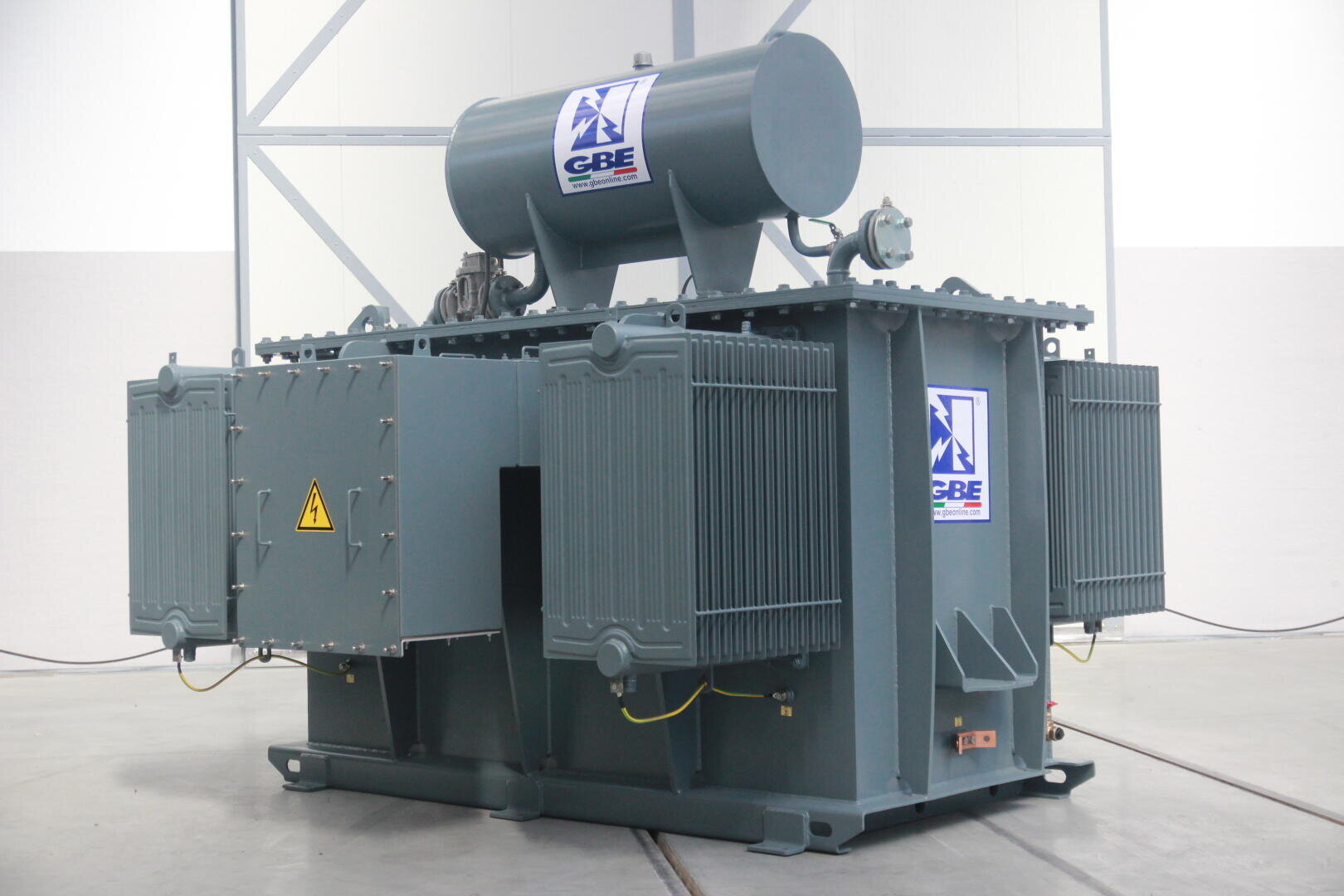 A large industrial electrical transformer with cooling fins and a cylindrical tank on top, located in a building on a concrete floor. The transformer is marked with GBE logos and a yellow high-voltage warning sign.