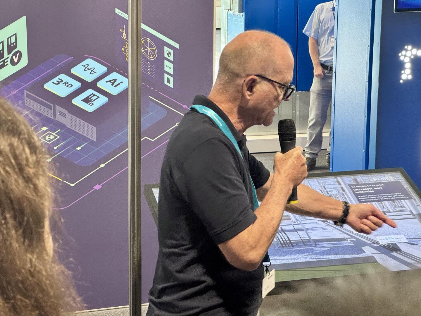 Ein Mann mit Brille hält ein Mikrofon in der Hand und deutet auf einen großen Touchscreen bei einer Technologieveranstaltung, während im Hintergrund Grafiken und Diagramme zum Thema Technologie zu sehen sind.