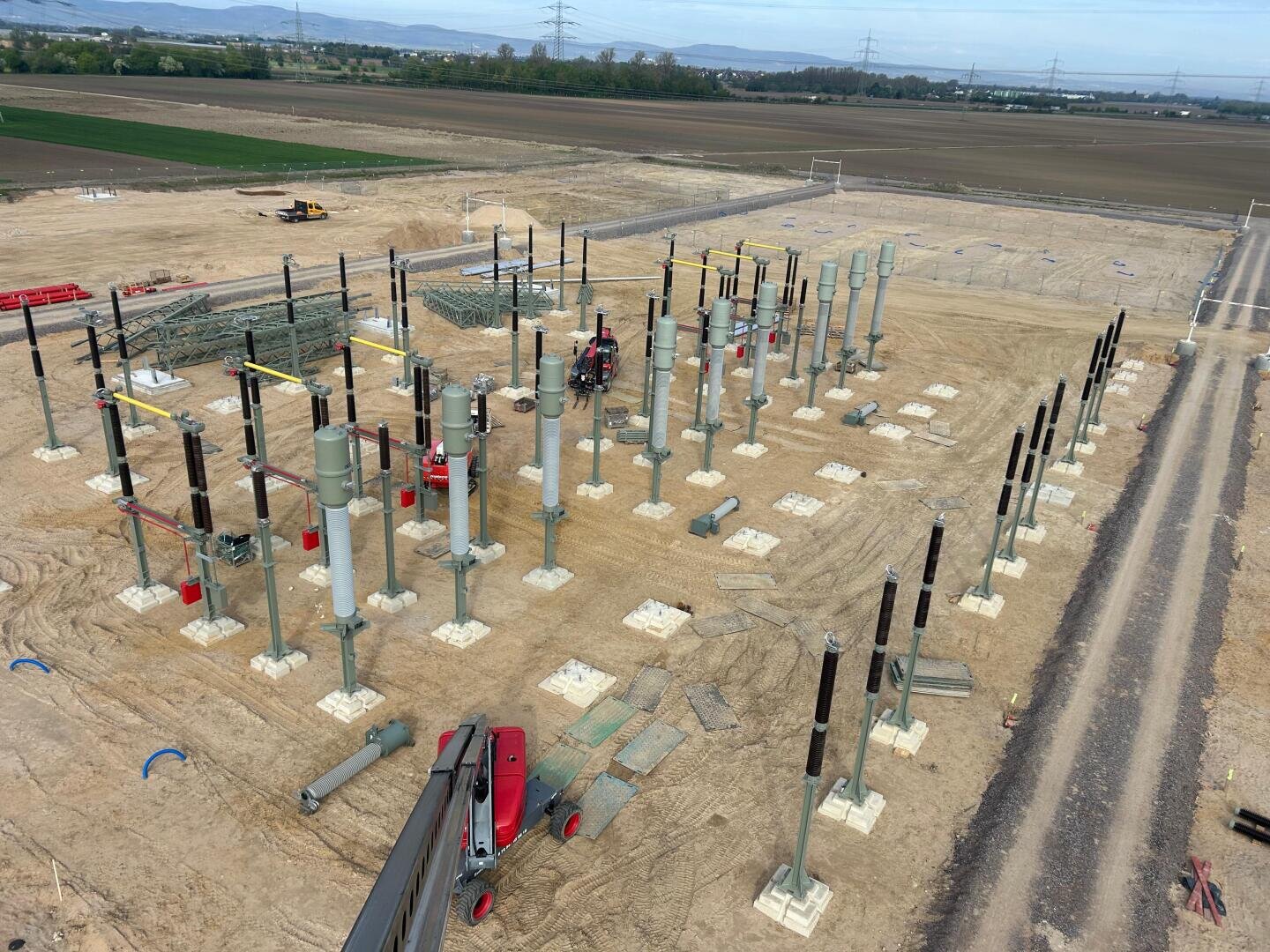 Luftaufnahme eines im Bau befindlichen Umspannwerks auf einem unbefestigten Grundstück mit zahlreichen Stahlsäulen und Isolatoren, umgeben von offenen Feldern und fernen Hügeln.