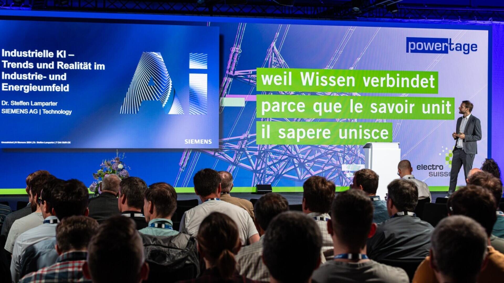 A speaker presents at a tech conference on industrial AI, facing an audience. The screen displays the Siemens logo and multilingual text about knowledge connecting people. Blue and green graphics fill the background.