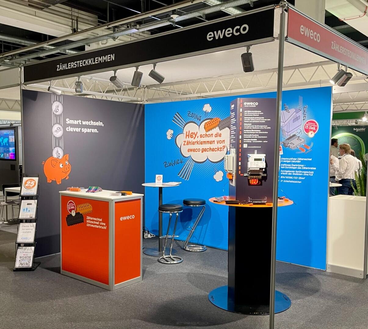 Der Messestand von eweco besticht durch farbenfrohe Grafiken, Informationsdisplays, eine Theke mit Stühlen, einen Prospektständer und eine Produktvitrine. Der Stand wirbt für Stromzähleranschlüsse unter dem Slogan „Smart wechseln, clever sparen“.