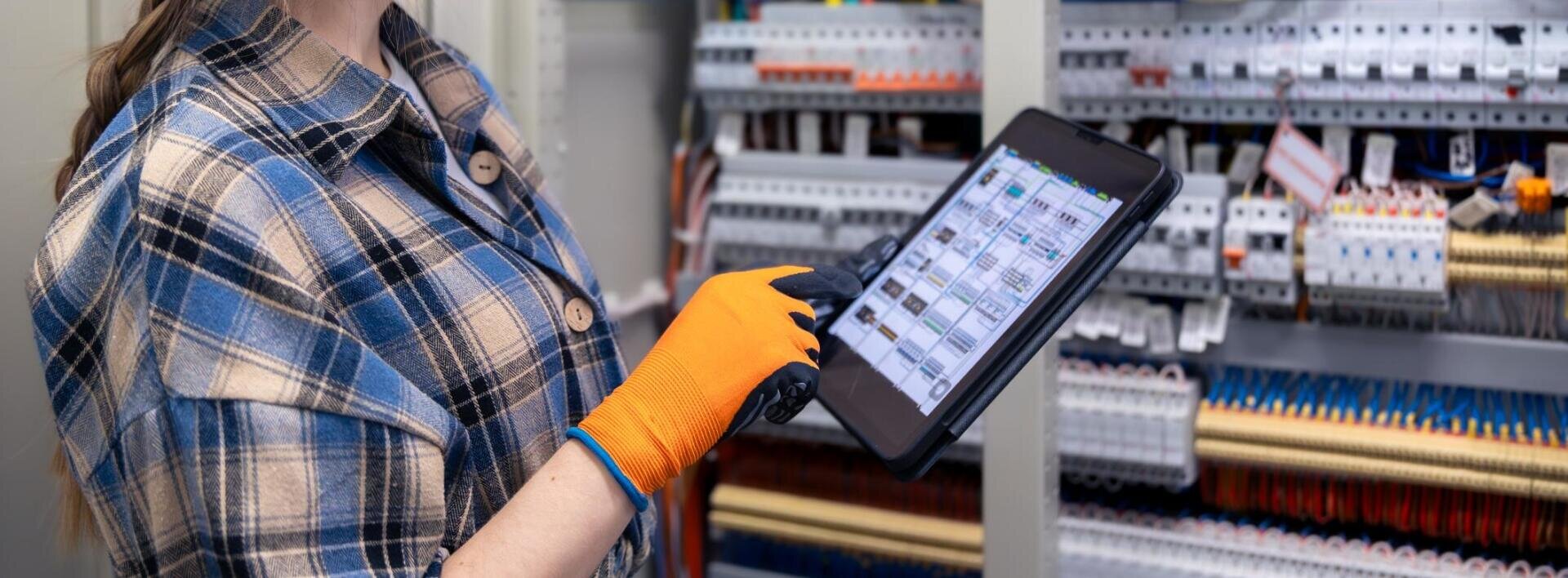 Eine Person mit orangefarbenen Handschuhen und kariertem Hemd hält ein Tablet in der Hand und prüft elektrische Schalttafeln mit Drähten und Schaltern in einer Industrieumgebung.