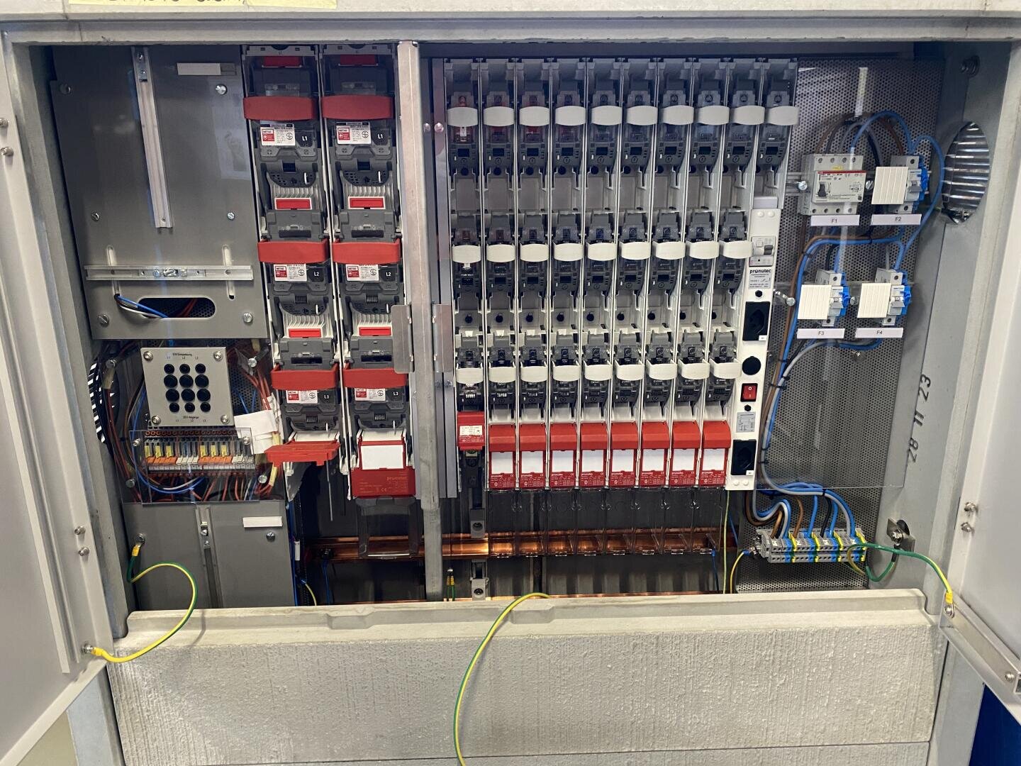 Offene elektrische Schalttafel mit mehreren Leistungsschaltern, Schaltern, Sicherungen, Kabeln und Klemmenblöcken, die übersichtlich in einem Metallgehäuse angeordnet sind. Die gelben und grünen Erdungsdrähte sind an der Unterseite sichtbar.