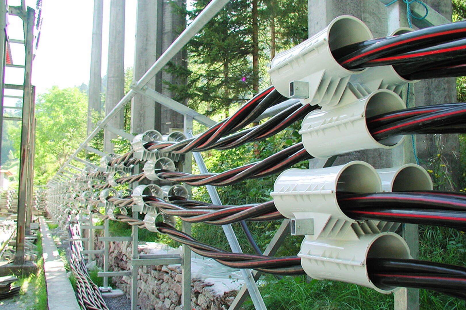 Mehrere Bündel großer schwarzer Stromkabel sind ordentlich angeordnet und werden von weißen Kunststoffklemmen an einem Metallgerüst im Freien gehalten, wobei im Hintergrund Grünpflanzen und Betonpfeiler zu sehen sind.