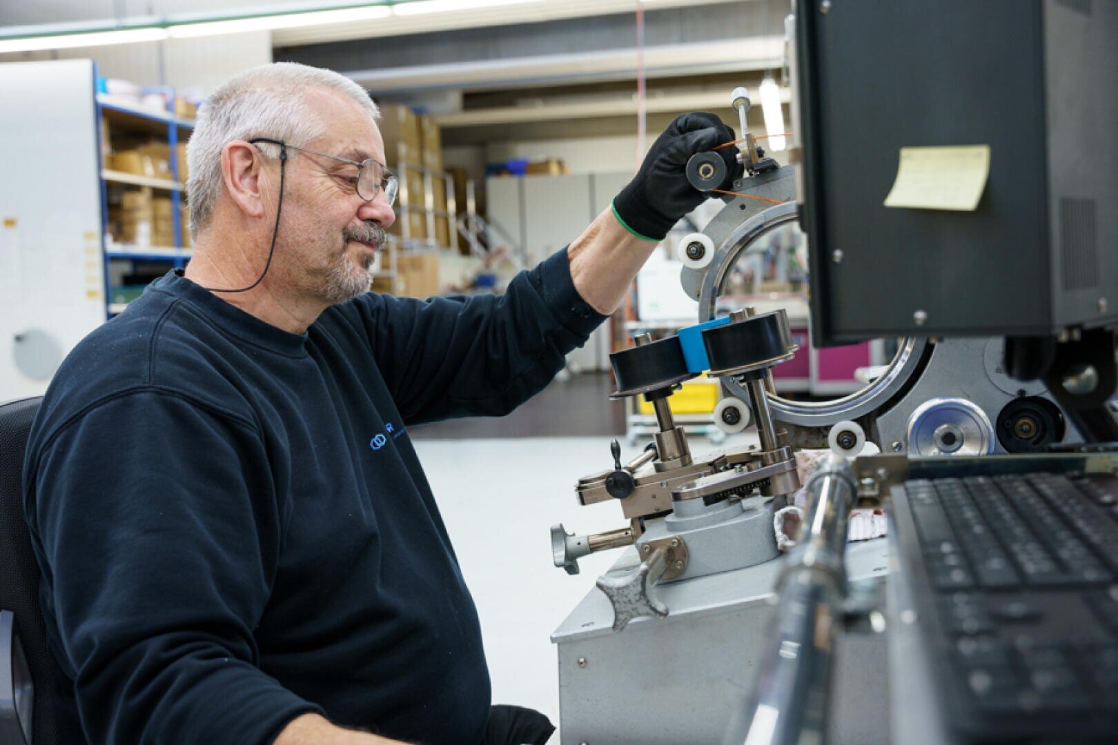 Ein Mann mit Brille und schwarzem Pullover bedient in einer Industriehalle eine Präzisionsmaschine und justiert ein kreisförmiges Bauteil, während er an einem Arbeitsplatz sitzt.