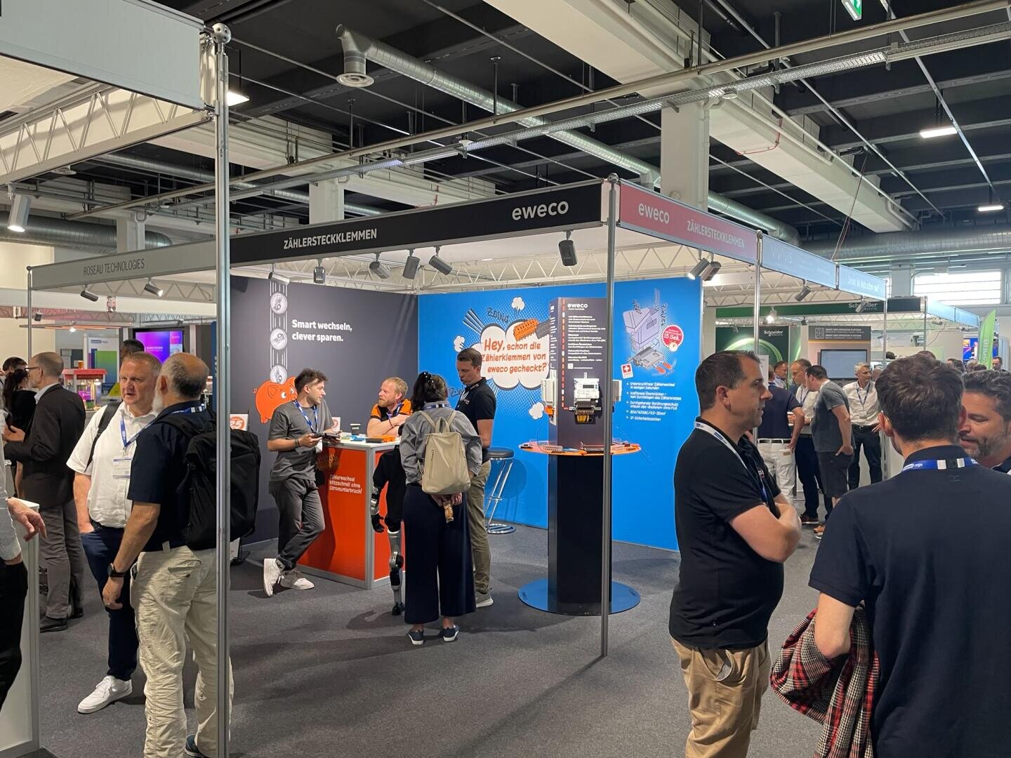 Ein belebter Messestand, an dem sich die Menschen unterhalten, herumstehen und miteinander interagieren. Der Stand ist mit dem eWECO-Logo, Informationsdisplays und digitalen Bildschirmen ausgestattet, in den Farben Blau und Orange gehalten, und mehrere Besucher tragen Namensschilder.