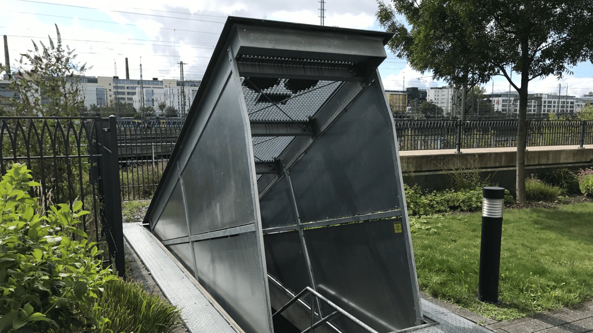Ein moderner, kantiger Metalleingang führt in den Untergrund, umgeben von Gras, Pflanzen und einem schwarzen Zaun. Im Hintergrund sind Gebäude, Bäume und Eisenbahnschienen bei teilweise bewölktem Himmel zu sehen.