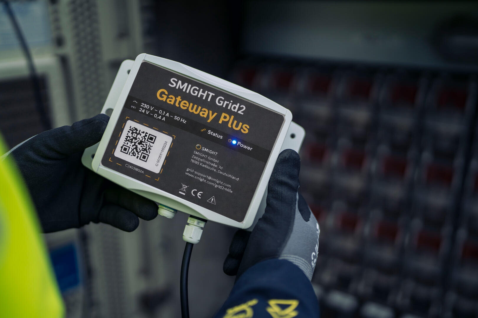 Eine Person mit Handschuhen hält ein SMIGHT Grid2 Gateway Plus-Gerät, das mit Kontrollleuchten und einem QR-Code auf dem Etikett versehen ist, vor ein elektrisches Gerät.