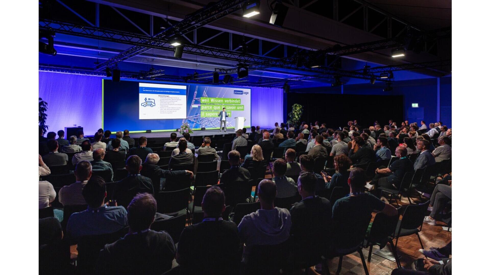 A large audience sits in rows facing a stage where two presenters stand in front of big screens displaying slides during a conference or seminar in a spacious, modern venue.