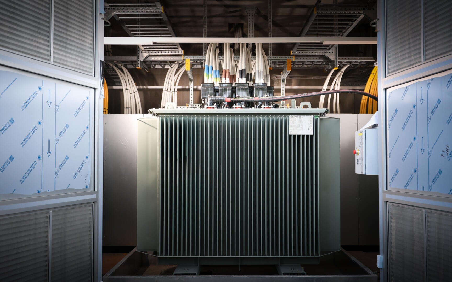Großer elektrischer Industrietransformator mit Kühlrippen, Kabeln und Verdrahtung an der Oberseite, in einem hell erleuchteten Raum mit Metallplatten und sichtbaren Leitungsrohren an der Decke im Hintergrund.