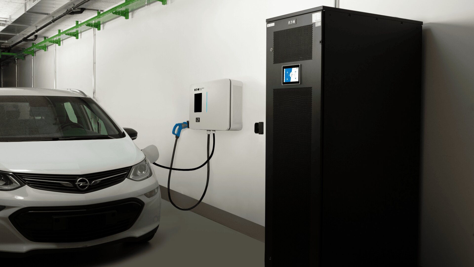 Ein weißes Opel-Elektroauto ist neben einer an der Wand montierten Ladestation geparkt, in der Nähe befindet sich ein großer schwarzer Schaltschrank in einer sauberen Inneneinrichtung.